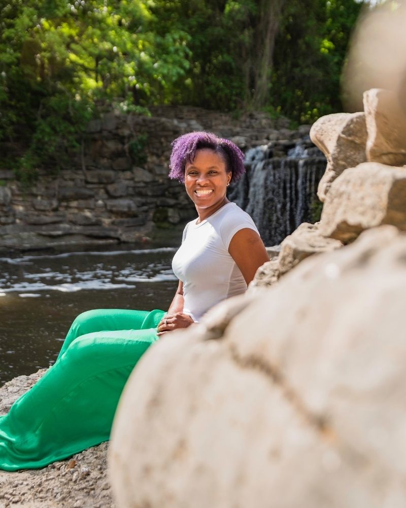 merissa-silence-about-page-photo Merissa Silence smiling while seated near a waterfall, wearing a white top and green pants